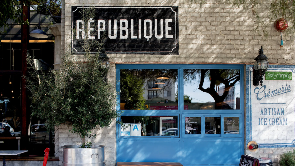 Republique is housed in a striking, historic building in Los Angeles, featuring elegant brickwork and arched windows that give the exterior a distinctly European feel. The grand entrance is adorned with a large wooden door and a marquee sign that reads "Republique" in bold letters, welcoming diners to this beloved restaurant.